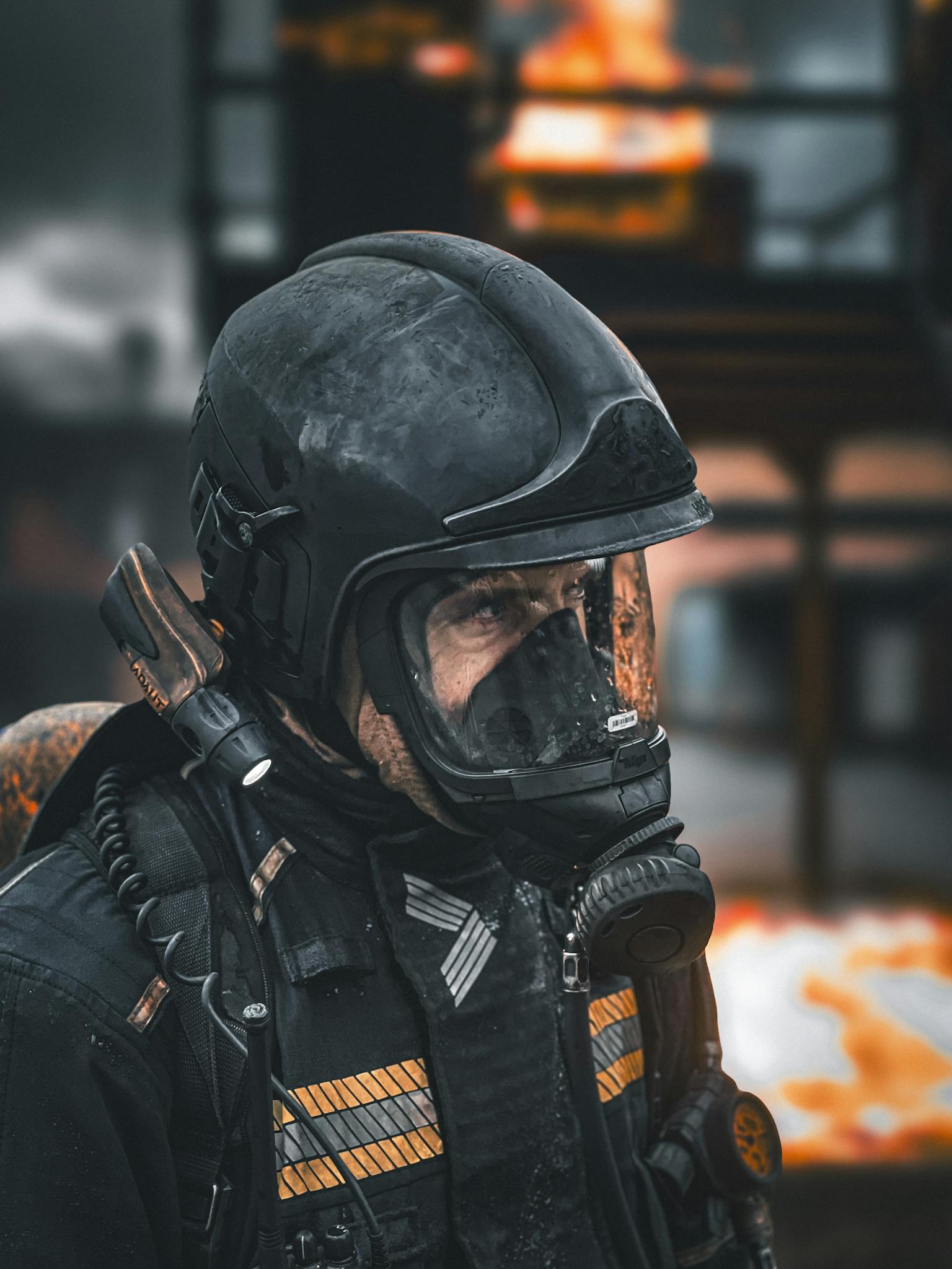 Firefighter with helmet and mask in action during blazing fire scene. Dark, intense atmosphere.